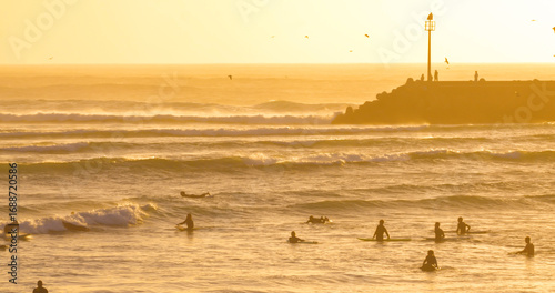 SILHOUETTE: Unrecognizable surfers wait and ride waves at a surf spot protected by a pier. Offshore wind and hazy atmosphere over the water and African coastline.