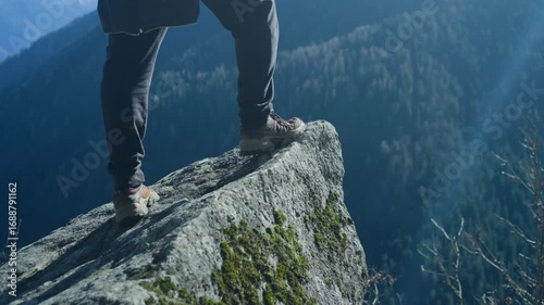 Tripod-level closeup of a hiker’s feet and legs stepping onto a rock over a mountain cliff, looking out at the scenic valley below.