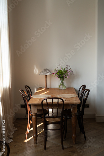 Spacious dining room interior with wooden farmhouse table and bentwood chairs in vintage style, vintage table lamp and vase with summer flowers. Aesthetic cozy interior with warm light