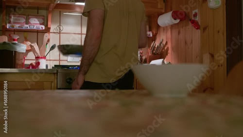 Tripod shot at countertop level of a man cooking with a pan on the stove in a wooden mountain cabin at night, seen from behind.