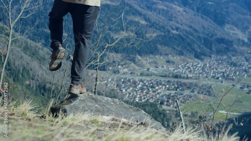 Tripod shot of a man balancing on one foot on a small rock in a mountain clearing, with grassy rocks and a blurred valley town in the background.