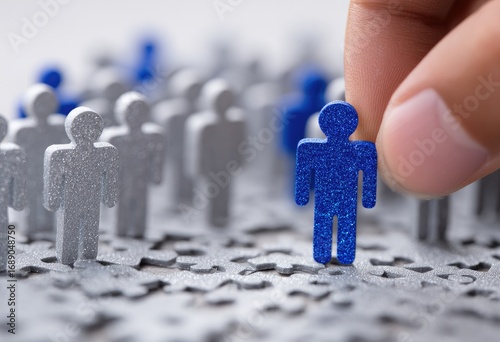 A hand places a blue figure among silver figures scattered on a surface with puzzle piece texture, focus on the chosen blue figure