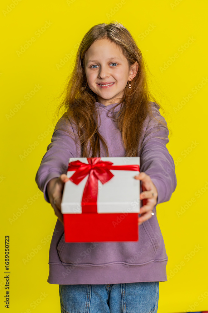 Fototapeta premium Lovely smiling young preteen child girl kid presenting birthday gift box stretches out hands, offer wrapped present career bonus, celebrating party. Children on studio yellow background. Vertical