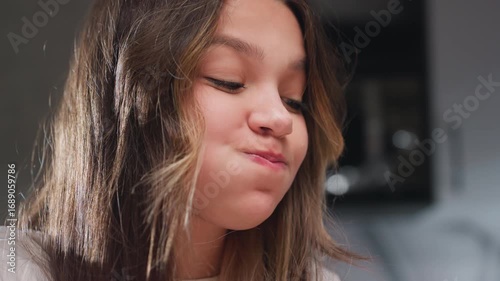 Close view schoolgirl with puffed cheeks while eating, playful turn to side glancing toward friend who laughs at cheeky face, warm light on hair, soft background bokeh, quiet kitchen moment