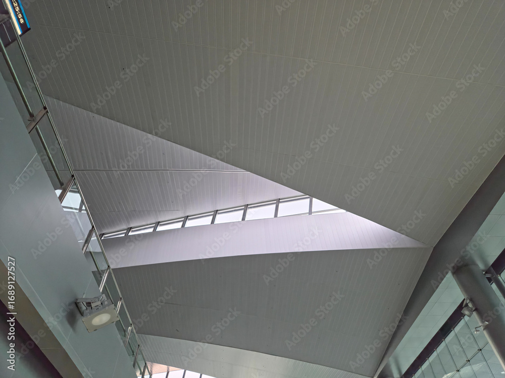 Obraz premium Upward view of a modern interior ceiling with slanted panels, geometric angles, a central window row, and a hanging light, emphasizing clean, dynamic design.