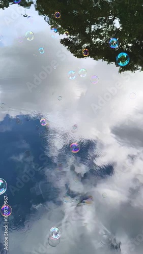 First-person view of blowing iridescent soap bubbles over a lake. Bubbles float and skip on water with sky reflections. Vertical handshot video, dreamy and playful scene.