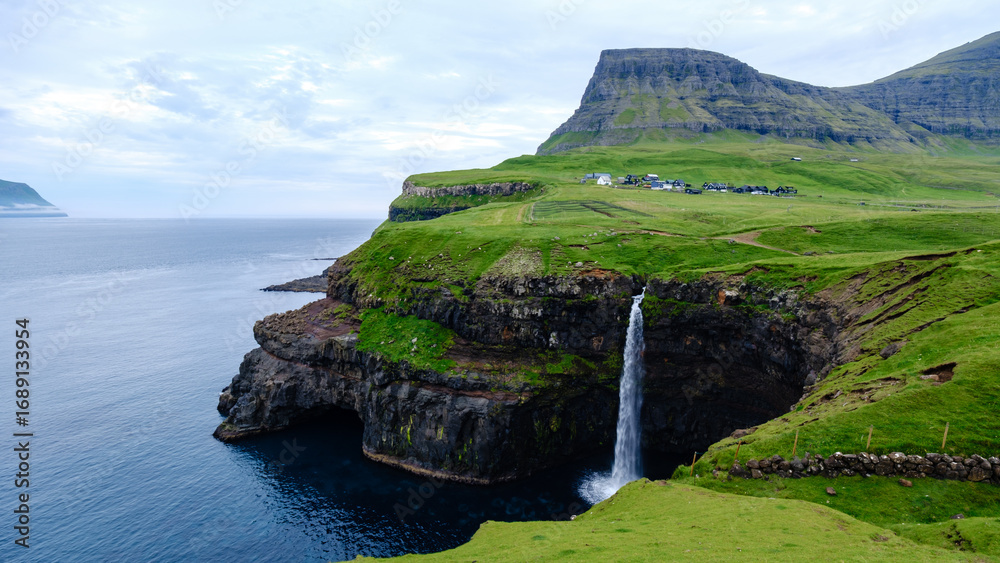 Naklejka premium waterfall tumbles from a green cliff into the Atlantic Ocean, portraying the rugged beauty of the Faroe Islands. Lush vegetation surrounds the area, Mulafossur waterfall near Gasadalur village