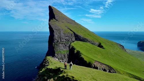 Two hikers stand atop dramatic cliffs in the Faroe Islands, surrounded by lush green landscapes and endless ocean. The blue skies create a perfect backdrop for adventure. Mylingur cliff hiking trail