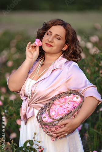 Romantic woman in light dress with curly hair sitting in blooming rose garden with picnic basket, enjoying summer nature outdoors, floral field lifestyle, relaxation and beauty in countryside scenery
