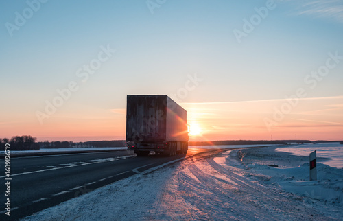 Semi truck driving along a snowy highway at sunset in winter
