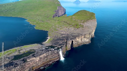 Majestic cliffs rise from the blue waters in the Faroe Islands, showcasing stunning natural beauty. A waterfall cascades down rocky formations, embodying the serene landscape, Sorvagsvatn lake, Vagar