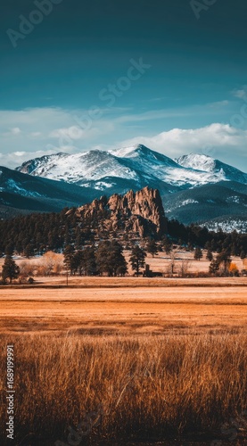 Scenic view of golden grasses in the foreground, a rocky hill in the midground and snow capped mountains against a teal sky in the background