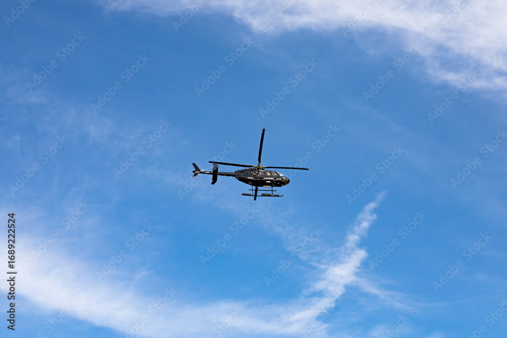 Fototapeta premium Black Helicopter in Flight Across Blue Sky with Scattered White Clouds