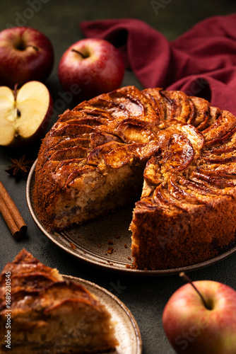Sliced ​​sponge apple pie with caramelized apples on green background next to burgundy kitchen textile napkin. Piece of pie on plate nearby. Apple harvest. Seasonal baking
