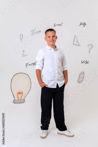 Thoughtful schoolboy in uniform sitting on wooden stool, student portrait with science and math formulas on white background, education and learning concept