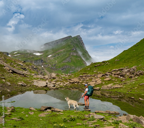 A person stands beside a calm mountain lake, enjoying a successful outdoor adventure with their dog. Lush green hills and rocky landscapes surround them under a bright blue sky.