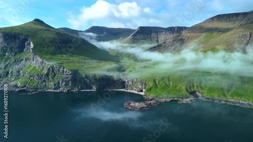 Discover the extraordinary beauty of the Faroe Islands with this aerial perspective. Lush green cliffs rise dramatically from the blue waters, while mist flows gently over the landscape.