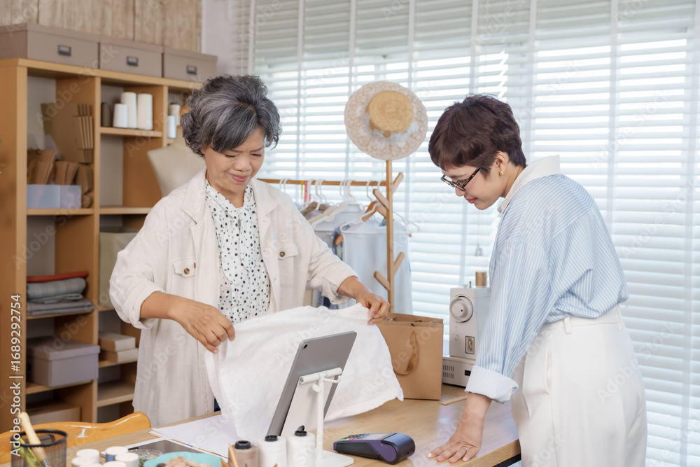 Obraz premium Senior asian seamstress folding handmade garment for adult female client at tailor shop counter with packaging bag, sewing tools and clothing rack, representing small business custom fashion
