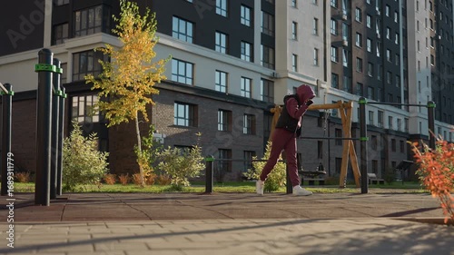Wallpaper Mural Fitness enthusiast holding pull up bar and stretching leg high beside metal bars in urban outdoor court under bright sun, wearing hoodie and sweatpants, modern residential background Torontodigital.ca