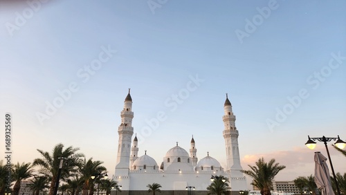 Quba Mosque, located in Medina, Saudi Arabia, is highlighted against a clear blue sky during sunset. The mosque features a prominent white facade with two minarets on either side.