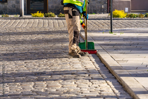 Worker in protective clothing sweeping cobblestone street with broom, standing near sidewalk curb, visible safety gloves and sturdy work shoes.