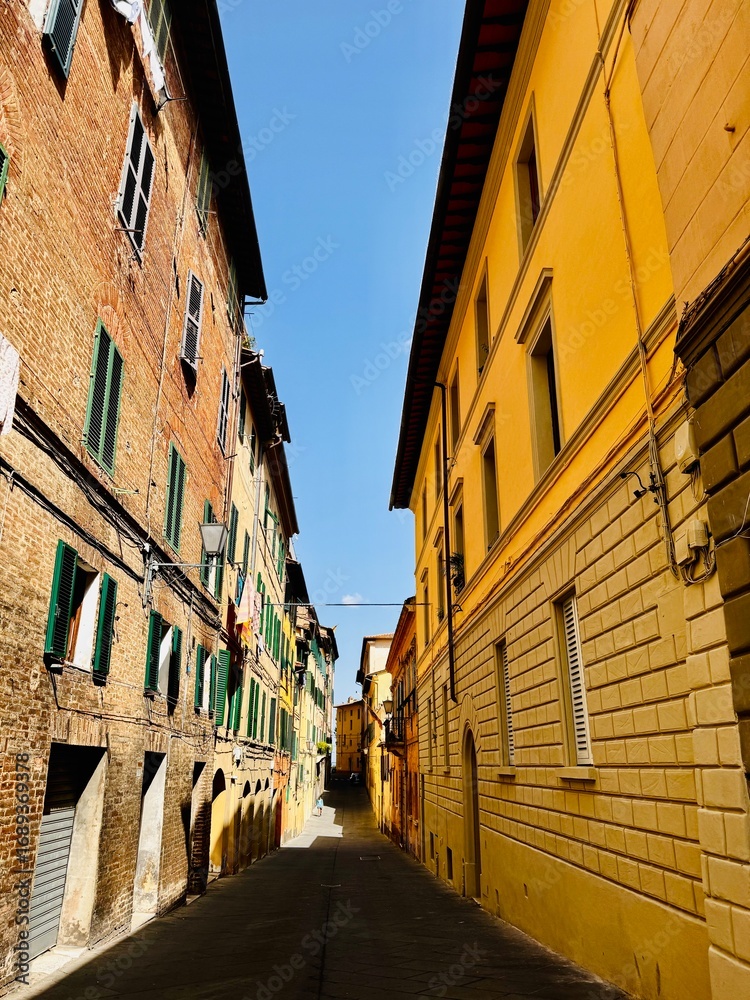 Naklejka premium Narrow cobblestone street lined with colorful historic buildings, featuring warm ochre and brick facades, green shutters, and laundry hanging above under a bright summer sky.