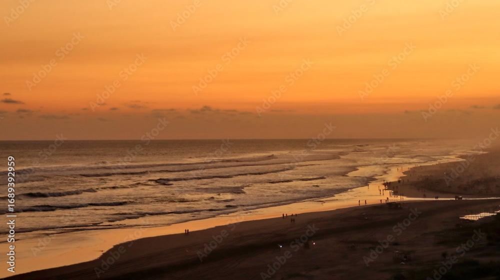 Fototapeta premium Scenic tropical beach sunset with golden sky, ocean waves and silhouettes of people along the coastline