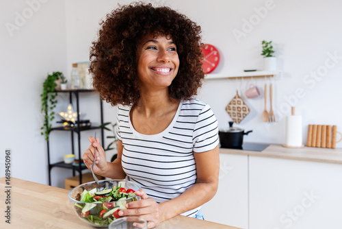 Wallpaper Mural Young happy woman with curly hair smiling and eating healthy fresh salad in her modern kitchen at home. Healthy eating habits lifestyle and food concept Torontodigital.ca