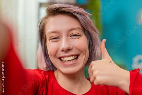 Young woman at home smiles at camera giving thumbs up showing approval and positive feedback during selfie. Girl with pastel hair and nose ring enjoys online connection indoors celebrating big success