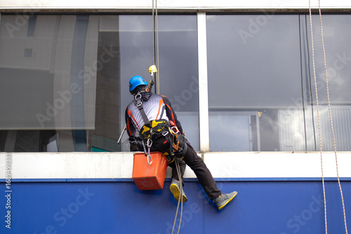 Rope access technician washing a building window with safety harness and helmet. Useful for window cleaning service, facade maintenance, industrial climbing, safety at height visuals.