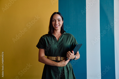 Portrait of smiling young businesswoman in smart casual with tablet standing against wall