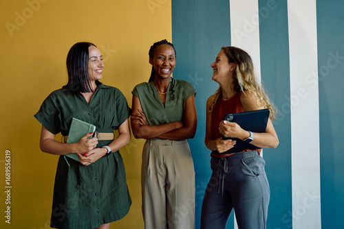Cheerful diverse businesswomen in smart casuals discussing against colorful wall in office