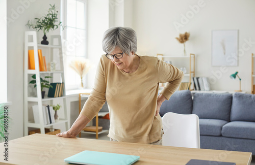 Fototapet Senior elderly woman standing at the desk in the living room at home touching her back suffering from backache