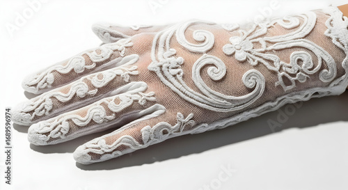 Close up of a hand wearing an ornate white lace glove with floral and swirl designs on a white background