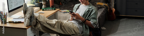 Teenage Caucasian boy sitting with legs on desk using smartphone while ignoring computer screen in bedroom, demonstrating gadget addiction and lack of focus on studies