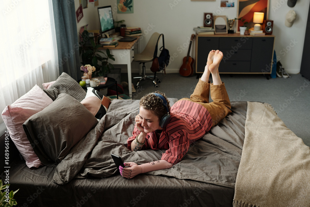 Obraz premium Young adult Caucasian woman lying on bed wearing headphones smiling while watching smartphone screen in bedroom showing signs of gadget addiction