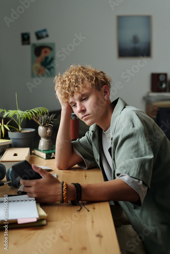 Fotografie Caucasian teenage boy sitting at desk holding smartphone with bored expression r