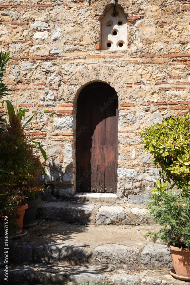 Fototapeta premium wooden door in a rustic stone wall, surrounded by green plants. steps lead up to the small archway, evoking a sense of history and secret gardens in a mediterranean setting