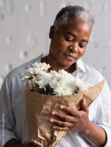 Mature African American woman with serene mood holding bouquet of white flowers against neutral textured background.