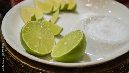 Slices of bright green limes are arranged on a white plate, accompanied by a small bowl of coarse salt, perfect for enhancing drinks or meals. The setting is cozy and inviting.