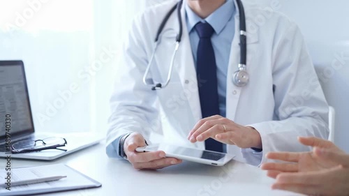 Doctor and a patient. The physician, wearing a white medical coat over a blue shirt and tie, is using tablet computer during a consultation in the clinic. Medicine
