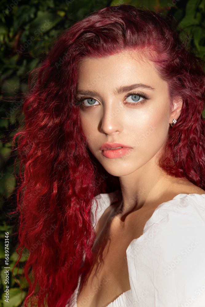 Fototapeta premium Beautiful young woman with long curly hair and gray-blue poses against the bush with hibiscus flowers. Portrait of a beautiful red-haired model with perfect skin and long eyelashes Real photo.