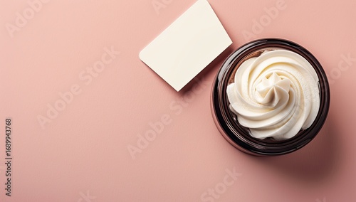 Creamy cosmetic product, elevated view.  A  whipped cream-like substance swirls inside a dark brown jar. A blank white card lies beside it.  Flat lay on a soft pink background
