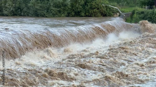 Close-up to the river swollen after heavy rainfall and flood water crashing through valley. The water flows fast from the high valley to the plain. General contest of a river in flood