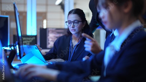 A team of data analysts and software engineers collaborates in a command center, reviewing real time analytics on a tablet while monitoring backend server performance and security logs.