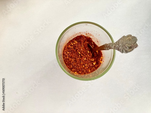 top view shot of a clear glass cup of red ้hot chili powder and a rose-patterned spoon sit on a white table with the copy space on the right side of frame