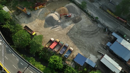Construction Site by Highway in July with Machinery and Piles of Material Surrounded by Greenery and Storage Trucks