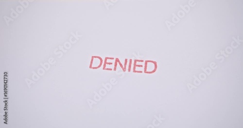 A real video frame showing a hand stamping the word ‘DENIED’ in red ink on a white paper document, symbolizing the official rejection of an application, loan, visa, claim, or request.