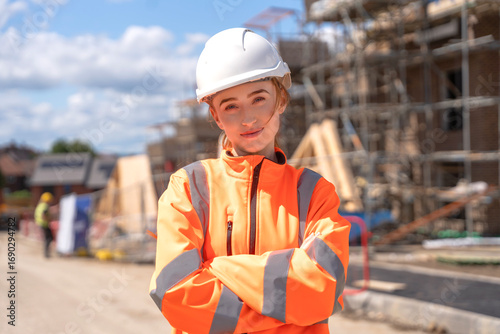 Young female construction worker poses confidently on site with safety gear during a sunny day with scaffolding in the background