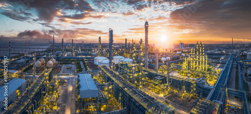 Spectacular aerial view of a massive, illuminated oil refinery complex at sunrise with the sun bursting through dramatic clouds.